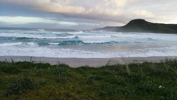A coastal landscape with waves crashing onto the shore, a grassy foreground, and a hill in the distance. - 5routes apartments connecting with nature (Vimianzo)