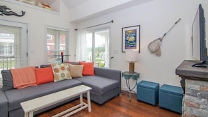 A living room with a grey sofa, a small table, and a stone fireplace. There are windows with blinds, a framed picture on the wall, and a guitar leaning against the wall. - Captain`s Quarters - 179 Leeward Way, Cottage #6 (Ottawa)