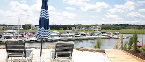 A beach area with two lounge chairs and a blue striped umbrella, overlooking a marina with boats and houses in the background.