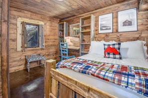 Camera da letto in una baita di legno, con un letto, una sedia, un tavolino, una finestra e mensole con cornici.