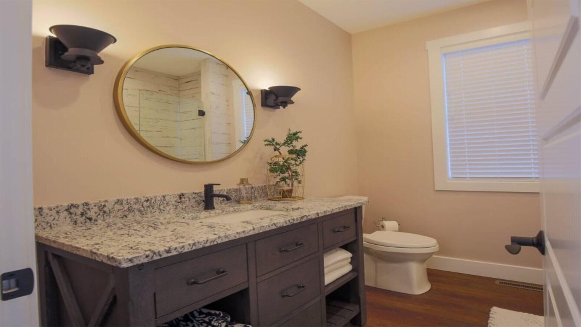 A bathroom with a granite countertop, a round mirror, a toilet, and a window with blinds.