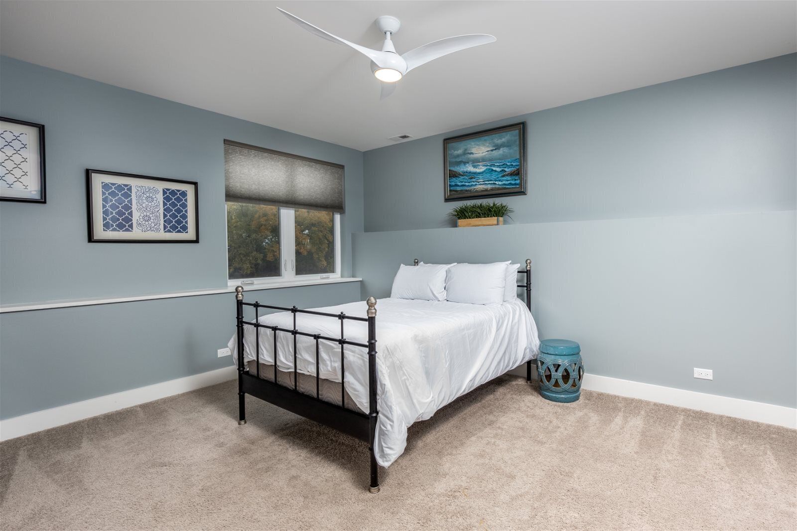 A bedroom with a bed, a ceiling fan, a window, and framed artwork on the walls.