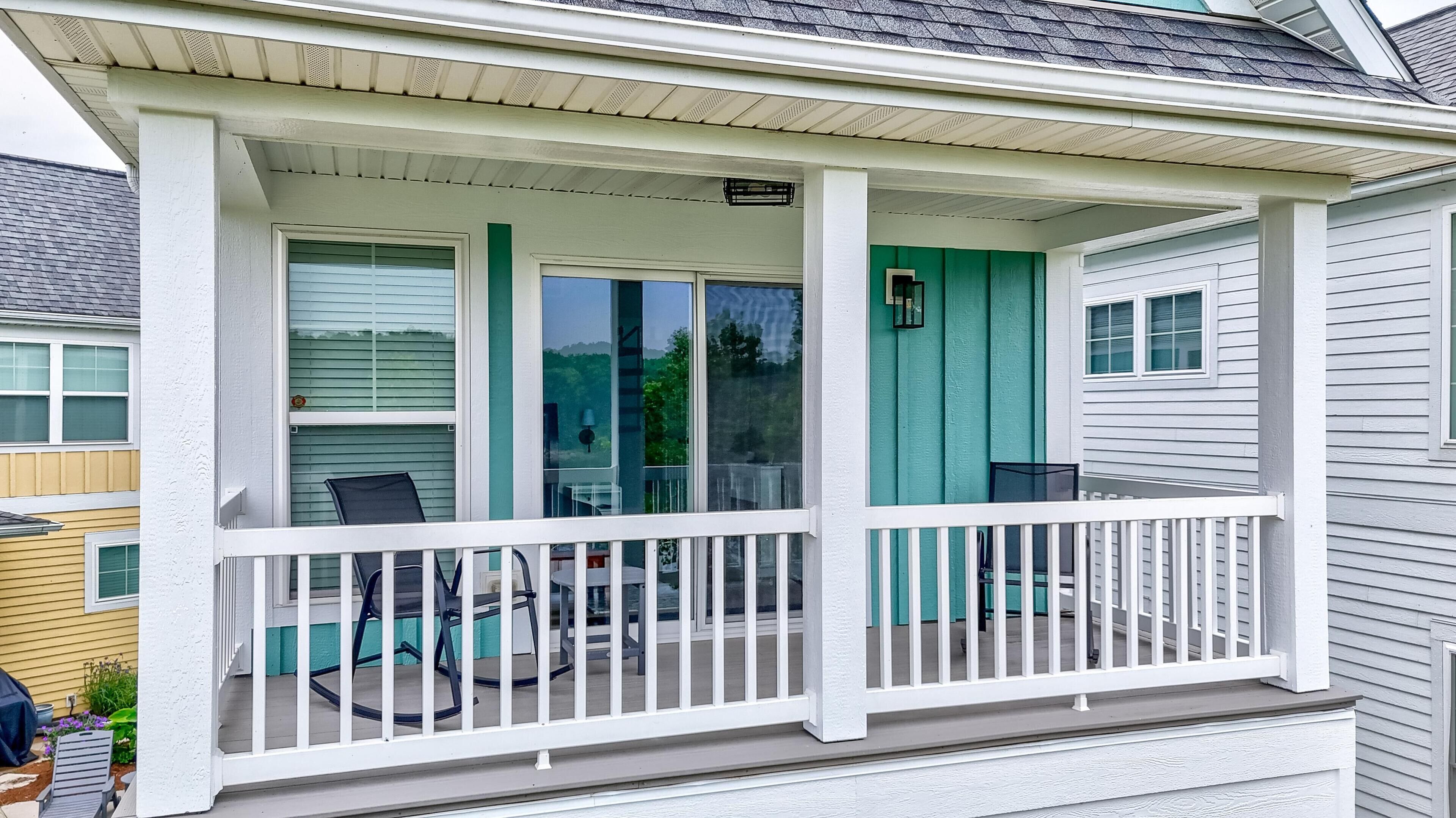 A covered porch with a white railing, a teal door, and a window with a view of trees.