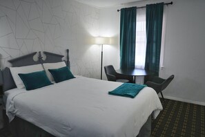 A hotel room with a bed, a desk, two chairs, a lamp, and a window with curtains.