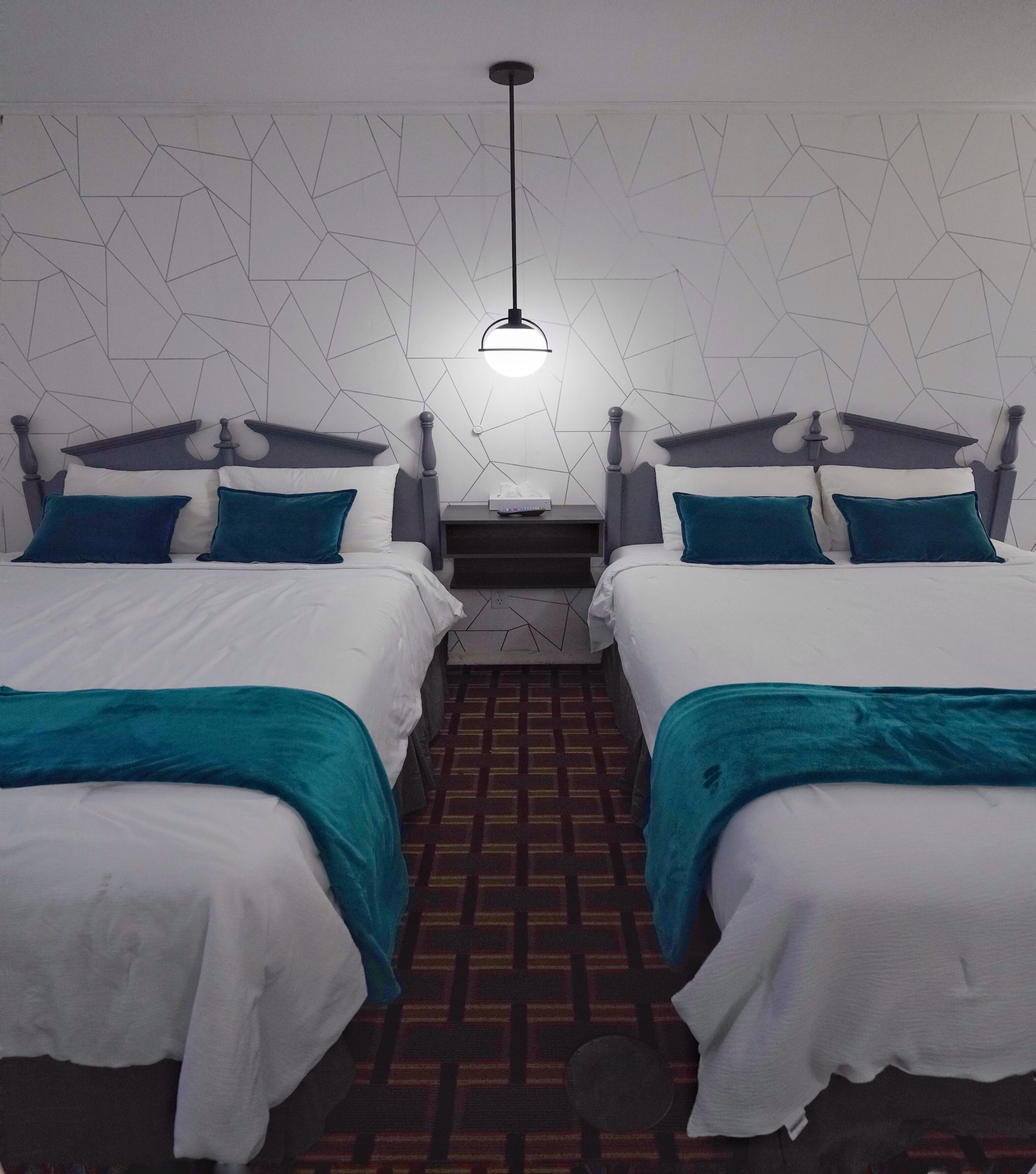 Une chambre d’hôtel avec deux lits, chacun doté d’une tête de lit, de draps blancs et de coussins décoratifs de couleur turquoise. La pièce est agrémentée d’un tapis à motifs et d’un mur orné d’un motif géométrique.