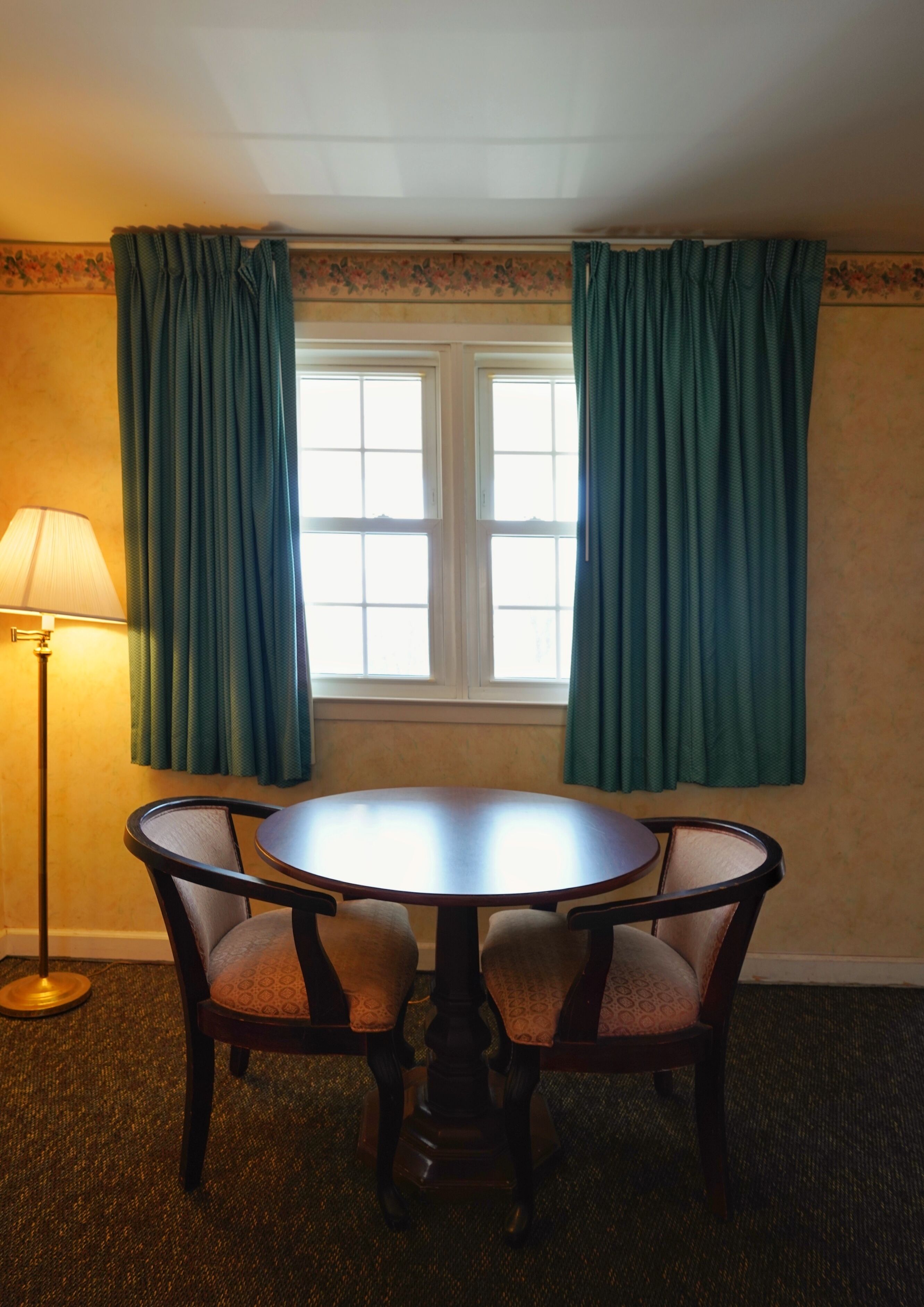 A room with a round table and two chairs, a floor lamp, and a window with curtains.