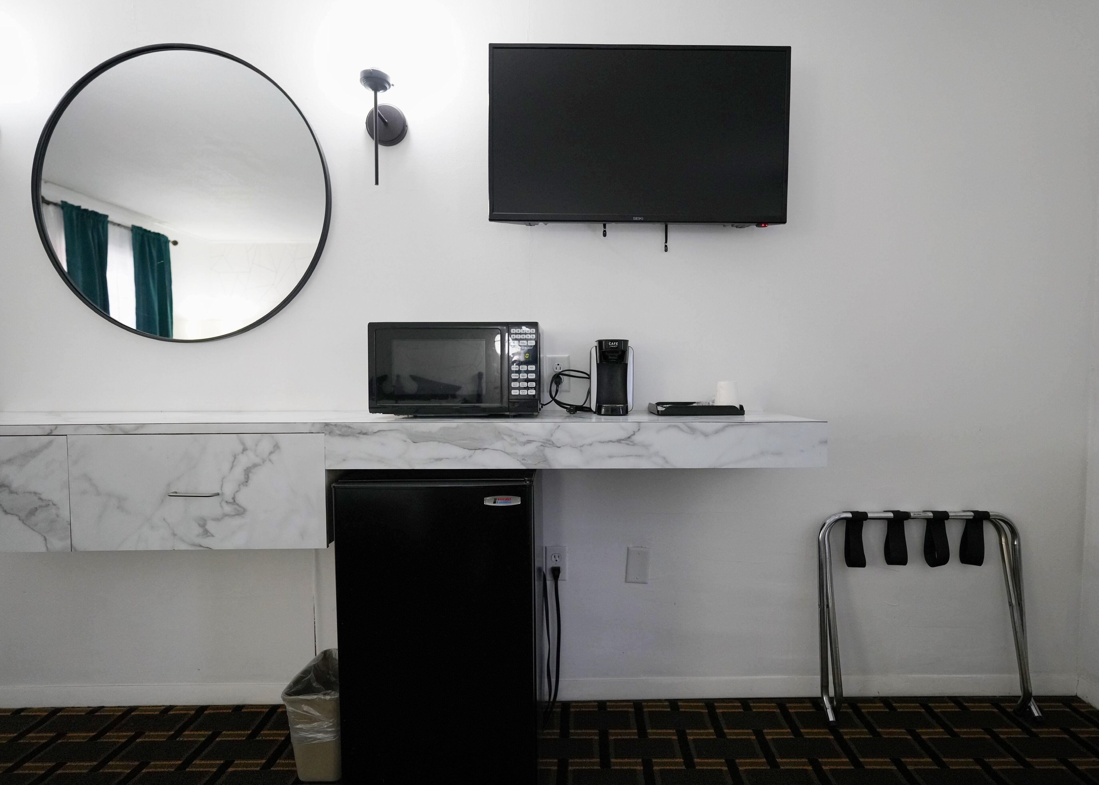 A compact hotel room with a white marble vanity, a black microwave, a coffee maker, a small refrigerator, a black trash bin, and a metal chair.