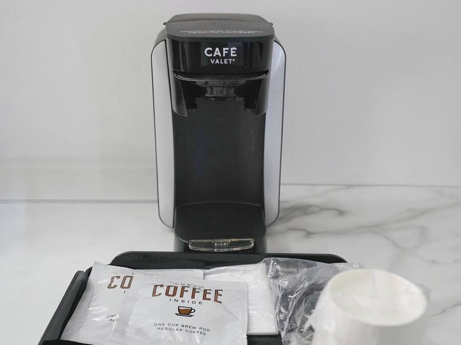 A coffee maker labeled "CAFE" on a tray with a coffee pod, a paper filter, and a plastic bag.