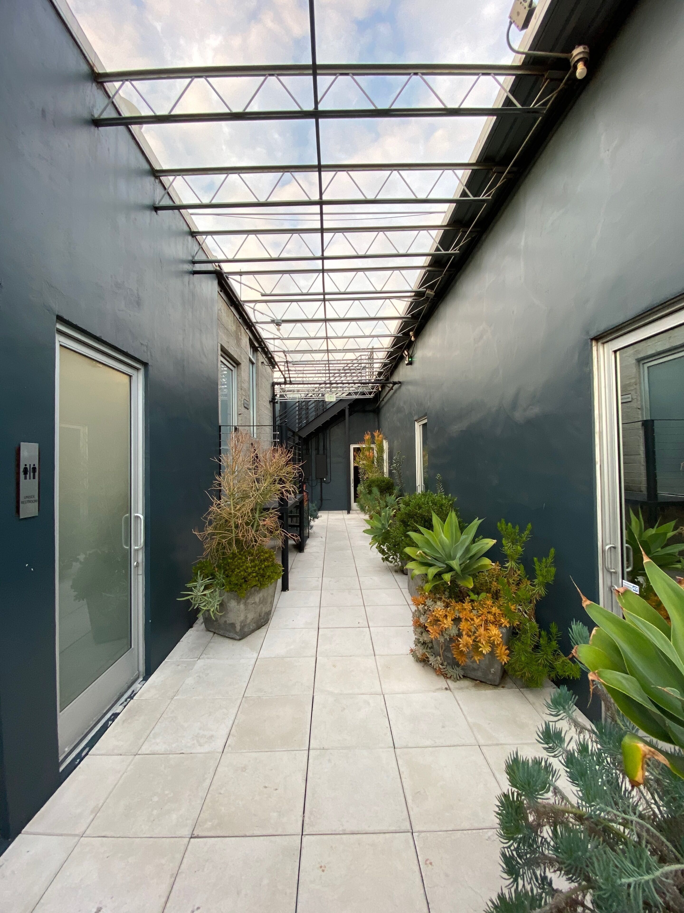 A corridor with a glass ceiling, potted plants, and a tiled floor.