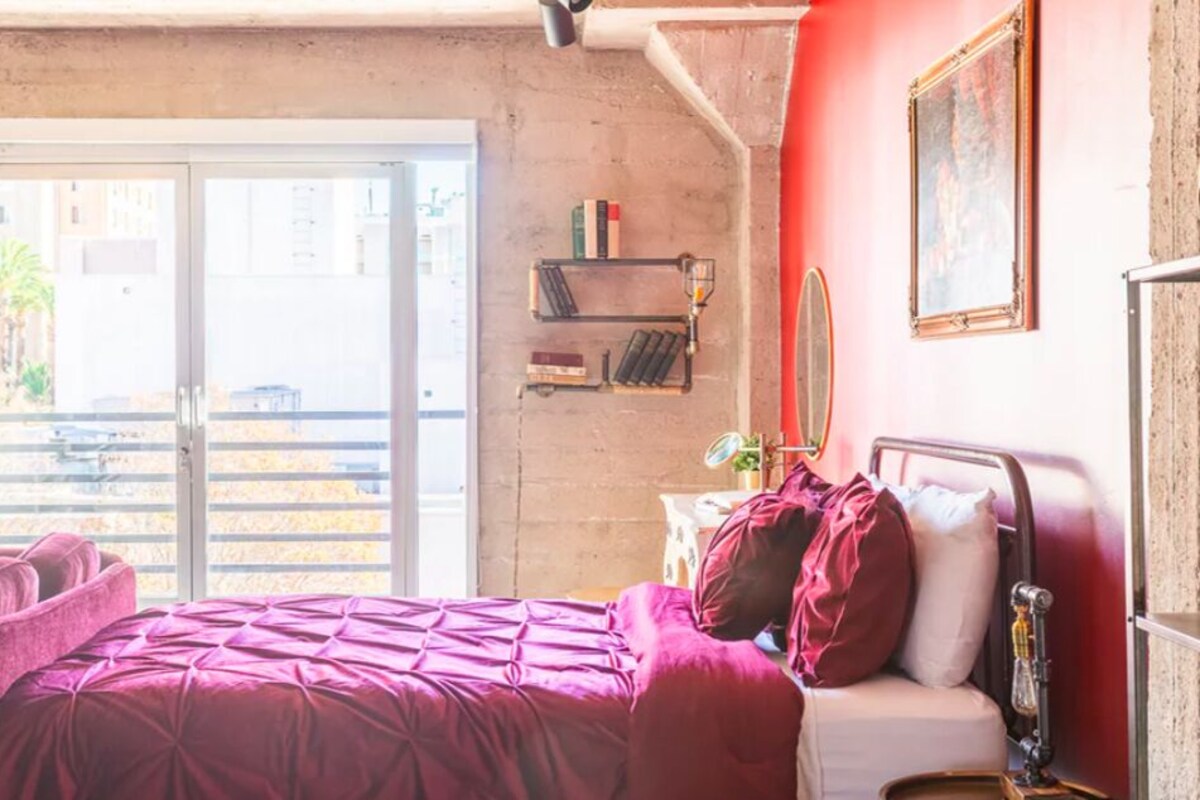 A bedroom with a bed, a chair, a bedside table, a mirror, and a window with a view of a building and palm trees.