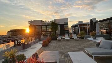 A rooftop terrace with outdoor seating, a view of the sunset, and a modern building in the background.