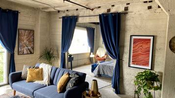 A room with a blue sofa, a coffee table, and a potted plant. There is a window with blue curtains and a painting on the wall.