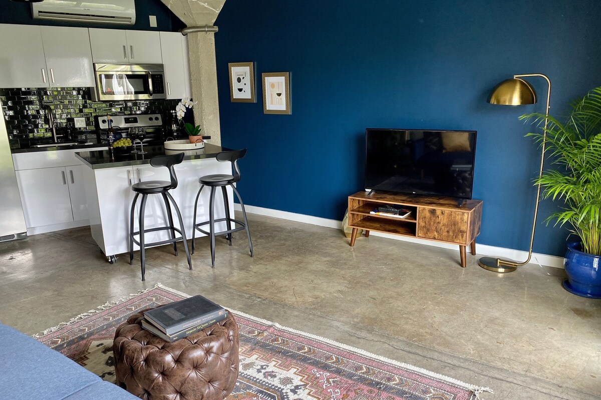 A modern kitchen with white cabinets, a microwave, and a coffee machine. There is a bar area with stools and a TV on a wooden stand. A patterned rug and a leather ottoman are on the floor.