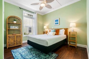 Ein Schlafzimmer mit einem Bett, einem Deckenventilator, einem Fenster mit Jalousien, einem Nachttisch mit Lampe und einem an der Wand befestigten Regal mit einer Pflanze.