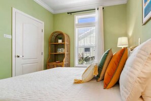 A bedroom with a bed, a window with blinds, a door, and a wicker shelf with a plant.