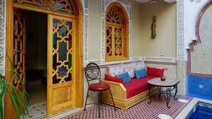 A room with a wooden door with intricate designs, a red sofa with blue and red cushions, a small round table with a red vase, and a patterned tiled floor. - Riad Shajara (Marrakech)