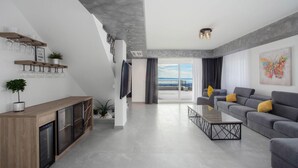 A modern living room with a grey sofa, a coffee table, and a TV mounted on the wall. There is a balcony with a view of the city.