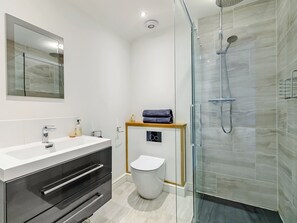 Et moderne badeværelse med et glasafskærmet brusebad, en hvid vask med et spejl over og en trækommode med skuffer.