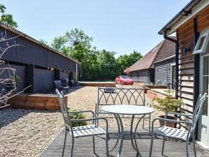 En terrasse med metalstole og et bord, en rød sportsvogn parkeret i baggrunden, og træbygninger med skrå tag.