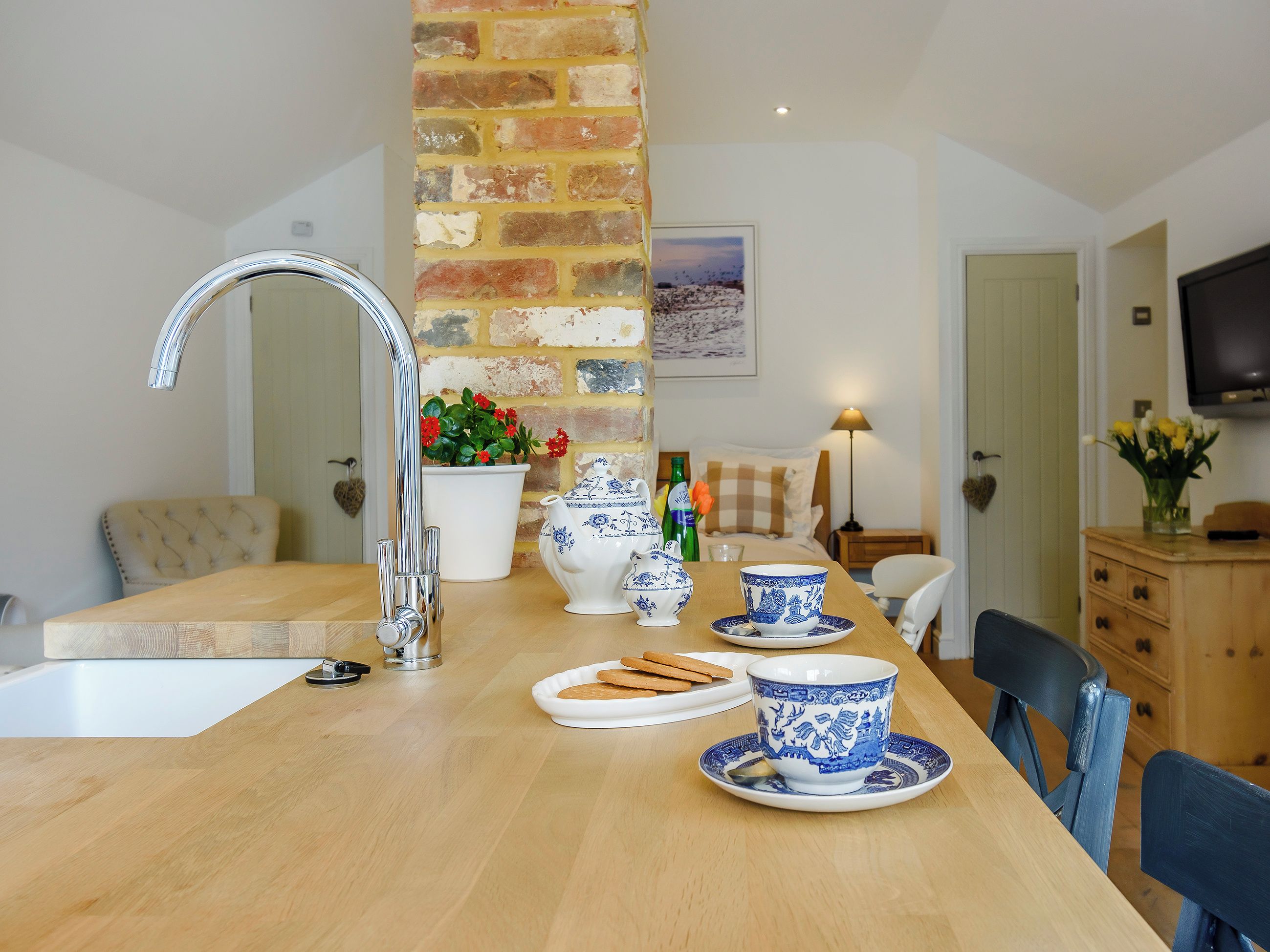 Een keuken met een houten tafel, een gootsteen en een bakstenen muur. Twee witte keramische bekers met blauwe motieven, een witte theepot en een witte schaal met koekjes.