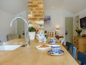 Een keuken met een houten tafel, een gootsteen en een bakstenen muur. Twee witte keramische bekers met blauwe motieven, een witte theepot en een witte schaal met koekjes.