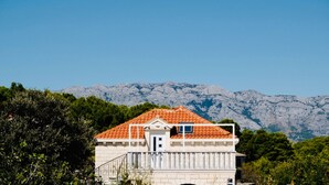 Un edificio con tetto in tegole rosse, un piccolo balcone e una porta. È circondato dal verde e dalle montagne sullo sfondo.