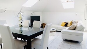 Ein modernes Wohnzimmer mit einem Glastisch, weißen Stühlen, einem weißen Sofa mit gemusterten Kissen und einem Flachbildfernseher.