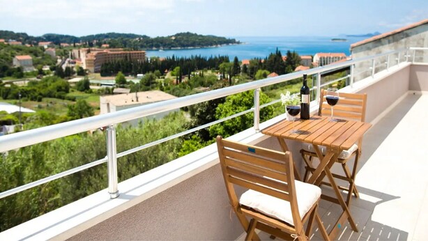 A balcony with a wooden table and chairs, a bottle of wine, and a glass of wine. The balcony overlooks a scenic view of a coastal area with buildings and greenery.