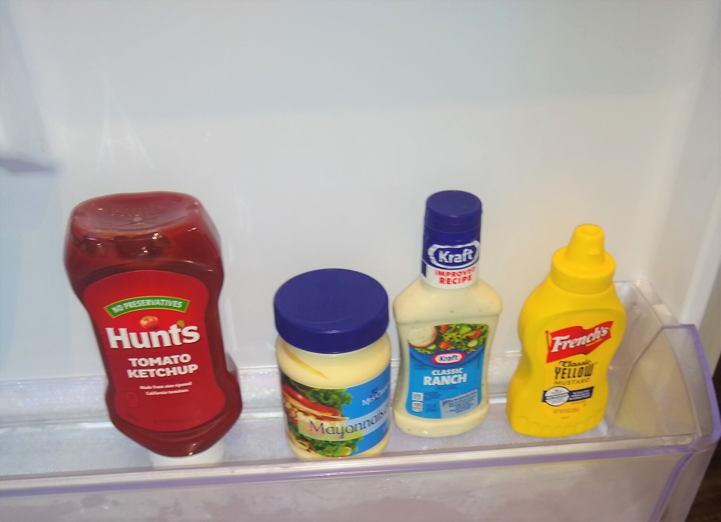 Une étagère de réfrigérateur contenant quatre bouteilles de condiments : du ketchup Hunts, de la mayonnaise Kraft, de la sauce ranch classique Kraft et de la moutarde jaune French's.