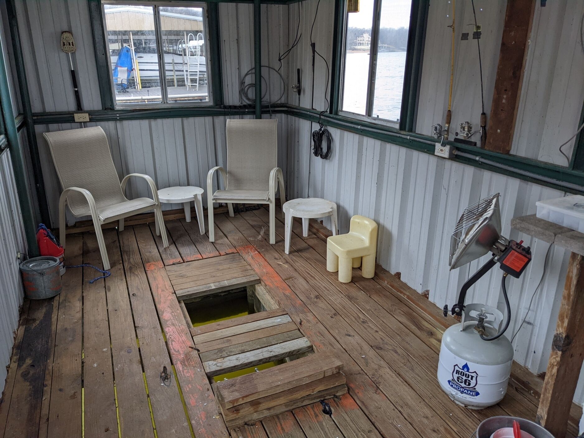 Een blokhut met een houten vloer, twee witte stoelen, een klein tafeltje, een ventilator en een propaantank.