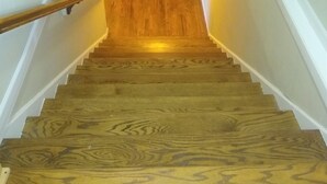 Eine Holztretreppe mit Handlauf, die zu einem Raum mit weißer Wand und Holzboden führt.