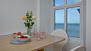 Ein Esstisch mit Obstschale, Blumen in einer Vase und zwei Gläsern rosafarbenen Getränks. Eine Uhr ist an der Wand.