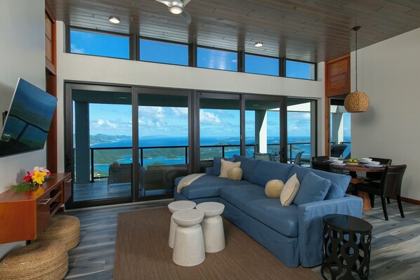 A modern living room with a large blue sectional sofa, a flat-screen TV, and a dining area with a view of the ocean.