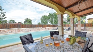 Ein überdachter Außenbereich mit Pool, Tisch mit Getränken und Obst sowie Gartenmöbeln.