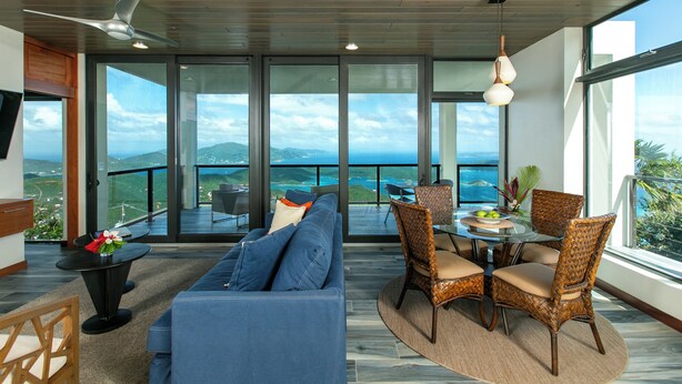 A modern living room with a blue sofa, dining table, wicker chairs, and a view of the ocean through large windows. - The Honeymoon Suite with Private Hot Tub (St. John)