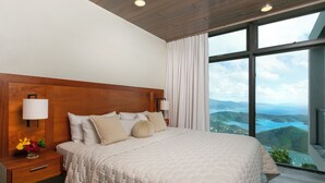 A bedroom with a large bed, wooden headboard, bedside lamps, and a view of a mountainous landscape through large windows. - The Honeymoon Suite with Private Hot Tub (St. John)