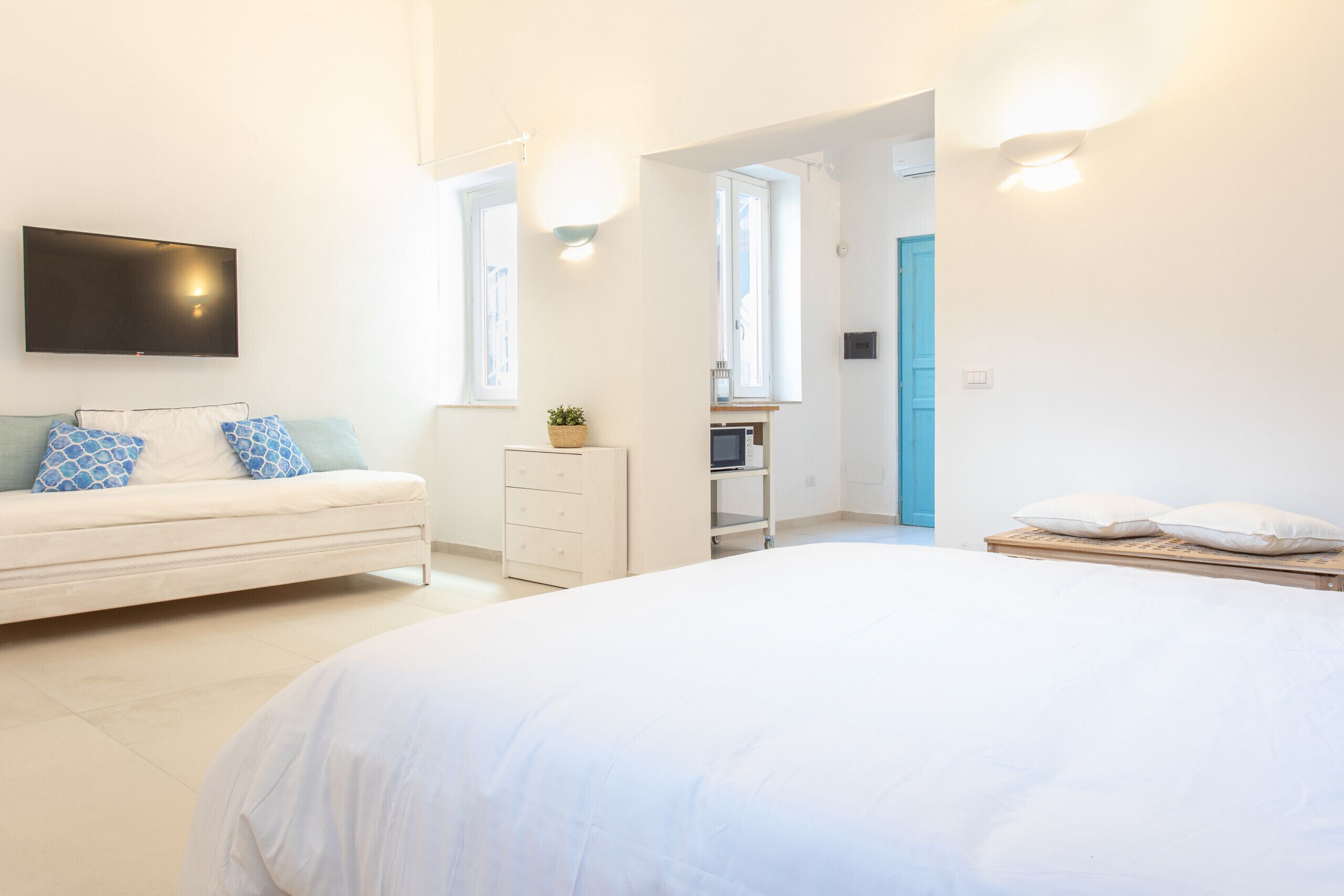 Een moderne slaapkamer met een groot bed, een aan de muur bevestigde televisie, een dressoir en een klein bureau met een plant.