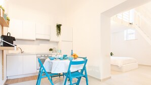 A modern kitchen with white cabinets, a dining table with blue chairs, and a staircase leading to a bedroom.