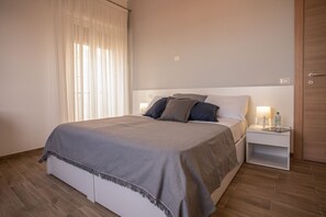 Un lit bien fait, recouvert d’une couverture grise, une table de chevet avec une lampe et une bouteille, et une porte en bois dans une chambre d’hôtel.