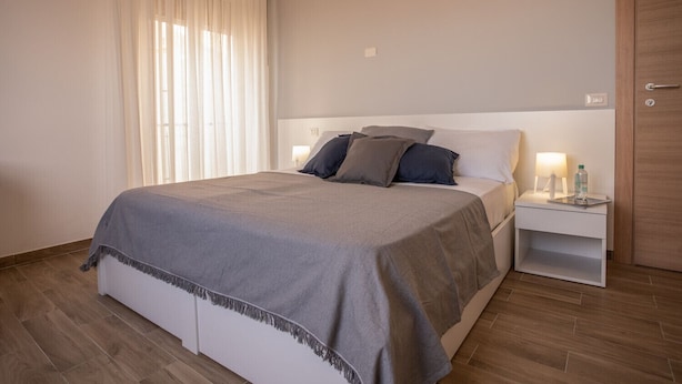 Un lit bien fait, recouvert d’une couverture grise, une table de chevet avec une lampe et une bouteille, et une porte en bois dans une chambre d’hôtel.