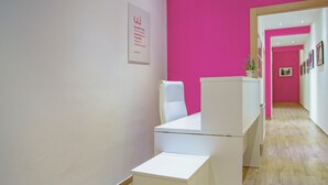 Un couloir avec un mur rose vif et des meubles blancs. Il y a un panneau sur le mur et une plante sur une table.