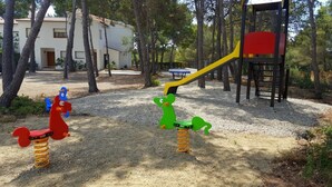 A playground with two swings and a slide, surrounded by trees and a building in the background. - Townhouses El Pinar   Rentalmar (Miami Platja)
