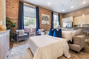 A modern hotel room with a brick wall, large windows, a bed with white and blue bedding, a grey sofa, and a kitchen area. - ✨GLAM MEETS DOWNTOWN HISTORIC CHARMER✨Spacious Corner Condo with Sleeper Sofa | The Stella (New Orleans)