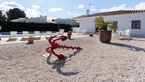A courtyard with a pool, white chairs, a red sculpture, and a white building with a tiled roof. - INNOUTHOME GARDEN, SWIMING-POOL,BEACH AT 150M,BBQ, AC AND WIFI_VIRGEN CINTA II (L'Ametlla de Mar)