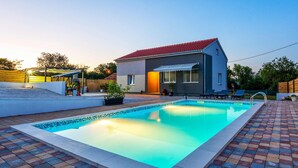 A swimming pool with a tiled deck, a covered patio, and a building with a red roof.