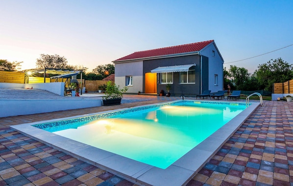 Una piscina con plataforma de azulejos, un porche cubierto y un edificio con techo rojo.