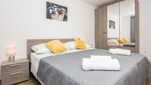 A neatly made bed with a grey quilt, two white pillows, and two yellow pillows. There is a bedside table with a lamp and a drawer. A large mirror reflects the room, and a framed picture hangs on the wall above the bed.