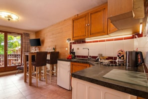 Una cocina con armarios de madera, un refrigerador pequeño, un comedor con sillas y un televisor montado en la pared.