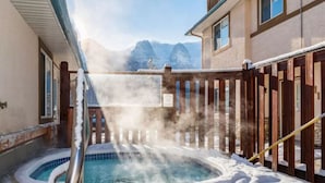 Uma banheira de hidromassagem rodeada por neve, com uma cerca de madeira e montanhas ao fundo.