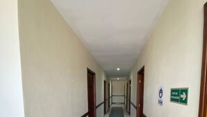 A long, narrow hallway with white walls and blue-tiled flooring. There are doors on both sides, and a green exit sign is visible on the right wall. - Hotel San Jose (Metapan)